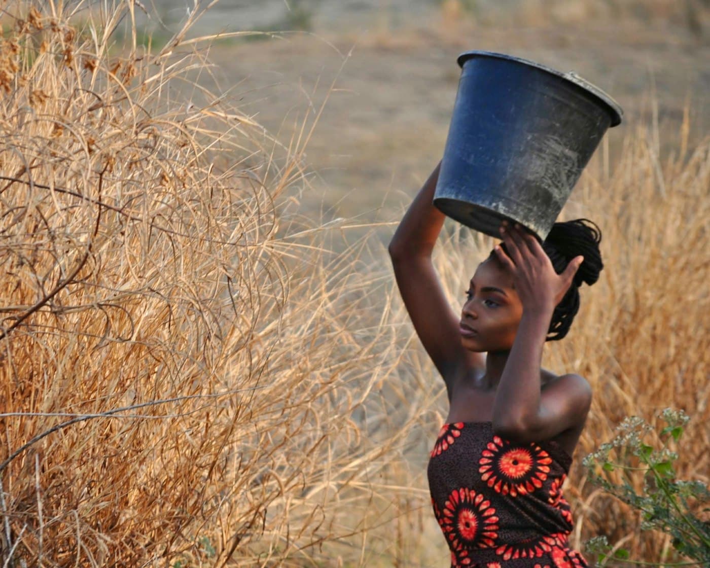 A young woman carrying a bucket on her head through dry grass