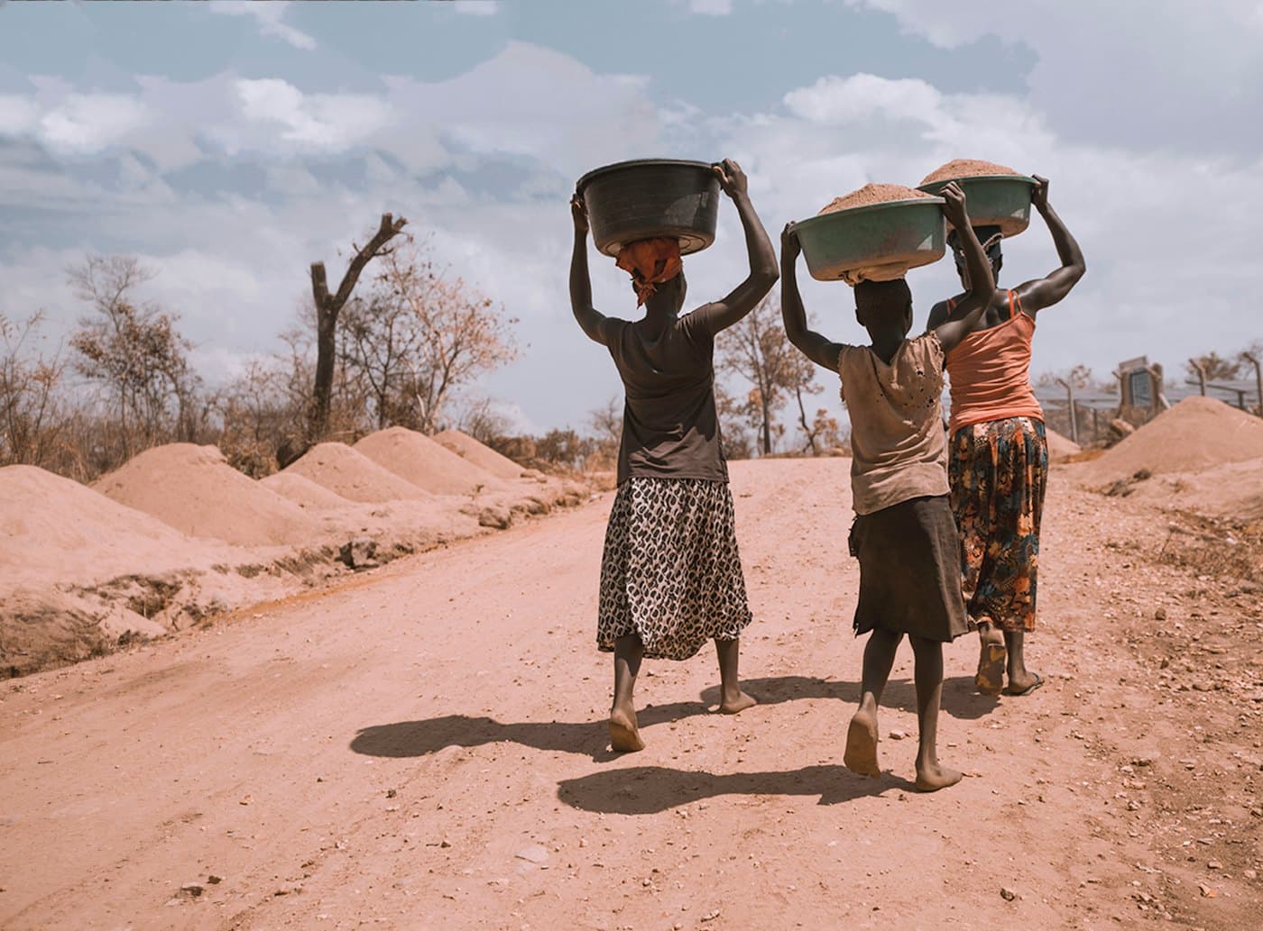 Three women carrying basins of water on their heads along a dusty road